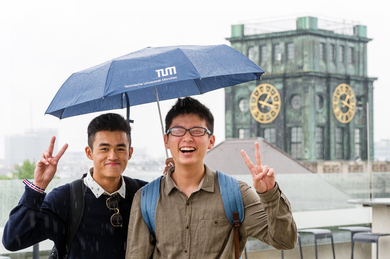 Zwei Studenten unter einem TUM Regenschirm, dahinter der Uhrenturm